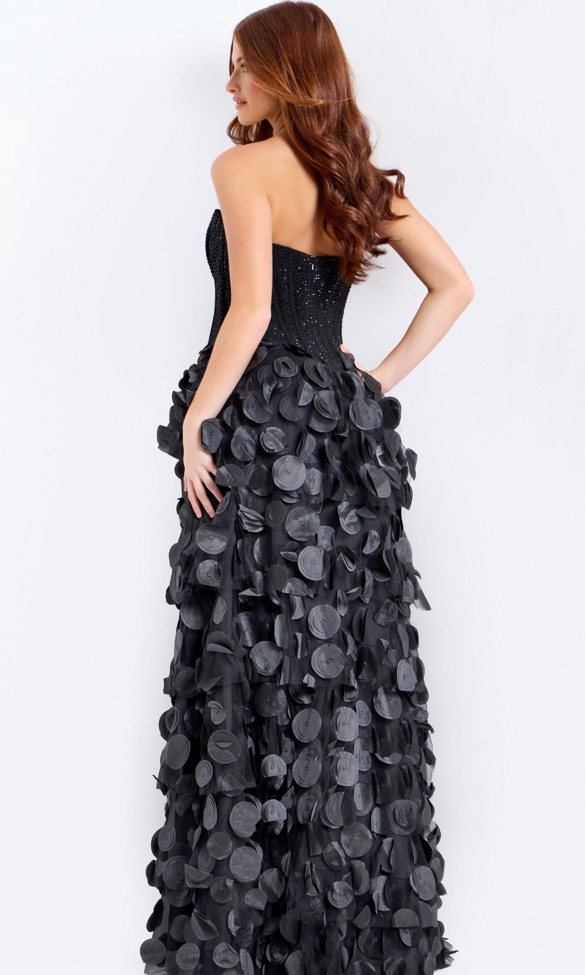 A woman with long, wavy hair models the Jovani 49384 Long Prom Dress, featuring a strapless beaded corset bodice and 3D petal A-line skirt, posing with her hand on her hip against a plain light background.